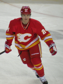 A hockey player in a red uniform with white and yellow stripes at the waist skates forward. His uniform has a white, stylized C logo on the chest and the number 24 on his arms.