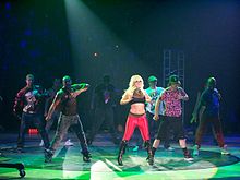 A blond woman female performer wearing a black and red outfit, dancing with a group of men.