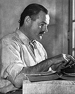 A black and white photo of Ernest Hemingway seated at a typewriter