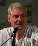 Photo en buste d'un homme avec les cheveux gris, portant une chemise blanche rayée et parlant dans micro devant lui.