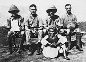 Photographie en noir et blanc représentant quatre hommes en uniformes militaires clairs, deux portant des casques en cuir (dont l'un a le bras en écharpe), assis devant un jeune garçon