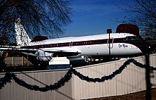 Photo couleur d'un avion aux couleurs blanches avec de fines bandes noires et rouges, entreposé au sein d'un enclos entouré d'un portail blanc.