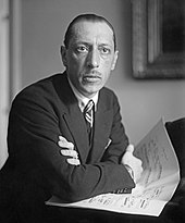 A man with short hair and glasses, wearing a dark suit and leaning against a piano