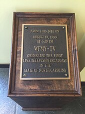 A wooden structure housing a historical marker plaque. Written on the plaque, "From this site on August 18, 1949, at 6:10 p.m., WFMY-TV originated the first live television broadcast in the state of North Carolina."