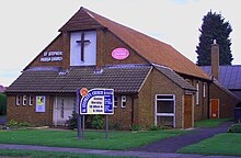Brick-faced church with a 45-degree tiled gable roof, with a large front-mounted cross on a white background, on grassy land, c. 2004.