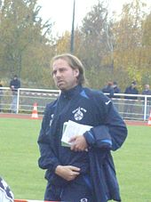 Photo d'un homme présent au bord d'un terrain de football. Il est habillé en survêtement de sport de couleur foncée avec le blason du club qui est en blanc. En arrière-plan, plusieurs personnes debout sont visibles et assistent à ce qui se passe sur le terrain.