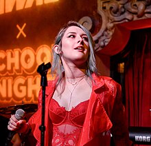 Mothica in a red outfit with shoulder-length hair holding a microphone on a stage