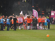 A crowd of men, some wearing grey suits and some wearing white shirts, navy shorts and white socks, celebrate raucously on a podium. An open bottle of champagne is visible in front of them, spiralling through the air as if somebody has thrown it.