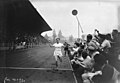 Photographie en noir et blanc d'un coureur à pied à l’arrivée d'une course, encouragé par des hommes qui crient.