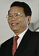 a smiling man with black hair, wearing glasses, and dressed in a suit with a red tie