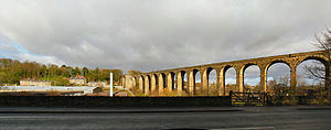 A curving stone viaduct bathed in sunshine on the right of the image. Houses and a small valley shown on the left.