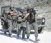 Four soldiers in green military attire walk in a line in front of a green military vehicle holding guns.