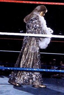 Photo en couleur d'une personne à cheveux longs sur un ring qui porte une longue robe à paillettes et des plumes autour du coup.