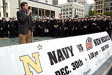 A man in a gray blazer and brown slacks speaks into a microphone behind a banner advertising the 2004 Emerald Bowl. Behind him are a few dozen young men in military uniforms.