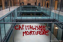 Tag à la peinture rouge : "Capitalisme mortifère" dans un hall propre et vitré.