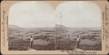 Man standing on a rock outcrop at a high elevation on a mountain with a view of the valley below. He has one hand in his pocket and is holding a hat with the other.