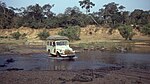 A photograph of a white vehicle with luggage on top driving across a river with green trees in the background all under a clear blue sky.