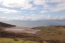 A sandy bay at left and an area of rough grassland at right lie next to a body of water in the middle distance. High hills under white clouds line the horizon.