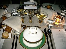 A set table with a white table cloth. There are many plates and glasses plus a menu visible on the table.