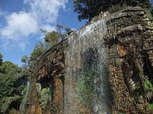 Vue de trois-quarts d'une cascade de quelques mètres de hauteur et de largeur dans un parc sous un ciel bleu.