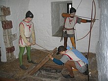 Photographie de plusieurs mannequins en habits médiévaux dans une salle aux murs blancs. L'un tire à l'arc par une meurtrière et les deux autres soulèvent une trappe en bois sur le sol.