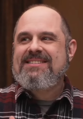 A 51 year-old man with a grey beard smiling to the left of the camera.