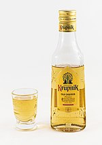 Kieliszek wypełniony cieczą o miodowym zabarwieniu. Obok niego stoi butelka nalewki marki Krupnik, z której przelano napój do kieliszka. Napis na butelce: Original Krupnik old liqueur Premium. Oryginalny Krupnik to legendarny smak i ponadczasowa jakość. Tradycyjny likier z najlepszych miodów i nalewek korzennych. Since 1846. Product of Poland. Starogard Gd. 1846. 38% vol. 200 ml.