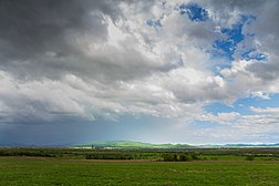 Photographie d'une plaine plate avec des champs au premier plan, des bois en second plan, et avec un ciel nuageux.