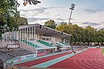 Tribüne mit grün-weiß-gestreiften Sitzbänken, von denen ein Stadion mit außenliegenden roten Laufbahnen beobachtet werden kann. Im Hintergrund Bäume und eine Beleuchtungsanlage.