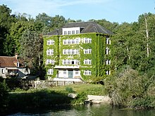 Photographie en couleurs d'un grand bâtiment à étages et à la façade envahie de végétation au-dessus d'un cours d'eau.