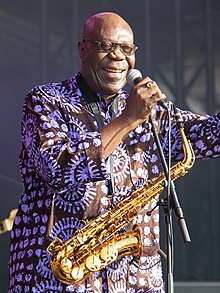 Manu Dibango de face avec un saxophone en bandoulière, tenant un micro à pied dans sa main droite, habillé d'une tenue colorée violette et noire.
