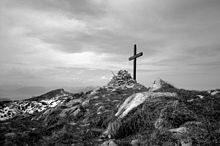 Photographie en noir en blanc d'une croix sommitale en bois et d'un cairn avec un ciel nuageux.