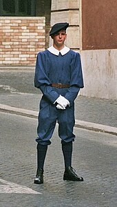 Frontale Farbfotografie eines Gardisten in dunkelblauer Uniform mit weissem Kragen und weissen Handschuhen. Er trägt schwarze Kniestrümpfe, schwarze Schuhe und schwarze Baskenmütze.