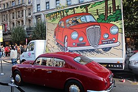 Photographie d'une voiture rouge stationnée devant un camion sur lequel est reproduit en grande taille une vignette de l'album.