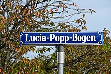 Blaues Straßenschild mit weißem Schriftzug Lucia-Popp-Bogen vor Herbstlaub