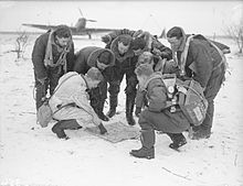 Photo noir et blanc d'un groupe d'hommes en tenues de vol regardant une carte posée sur un sol enneigé. Un avion est visible en fond.