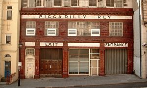 Side elevation of station with the typical red-tiled facade. A pale tiled band with the words "Piccadilly Railway" runs across the top of the second storey and "Exit" and "Entrance" are displayed above shuttered doorways.