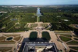 Farbfotografie in der Obersicht einer riesigen Parkanlage mit viel Wald und einer angelegten rechteckigen Wasserfläche im Hintergrund. Im Vordergrund sind breite Wege von ornamentalen Blumenbeeten unterbrochen. Am unteren Bildrand ist teilweise ein Schloss zu sehen.