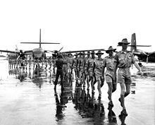 Des hommes en uniforme marchant sur un aérodrome avec deux avions de transport bimoteurs en arrière-plan.