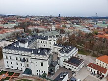 Farbfotografie in der Obersicht von einem weißen Schloss mit Seitenflügeln und schwarzem Dach. Links ist eine Kuppel und rechts ist ein gläserner Außenfahrstuhl. Im Hintergrund ist die Stadt mit Kirchen und einem Fernsehturm in der rechten oberen Ecke.