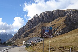 Pass-Straße in Fahrtrichtung Canazei mit der Talstation der Sass-Pordoi-Seilbahn. Links im Hintergrund die Langkofelgruppe.