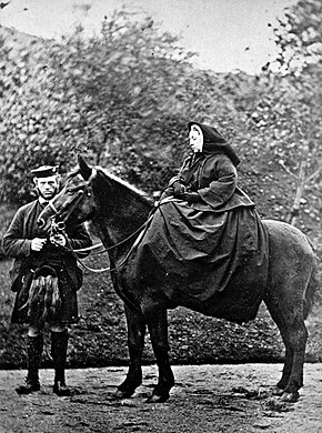 A woman, severely dressed in black, seated on a horse with a man standing by its head.