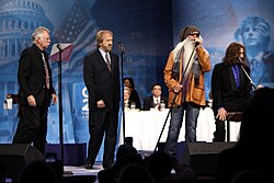 Four men standing at microphones on a stage. Three are wearing black suits. The fourth is wearing a tan jacket, dark glasses and jeans and has an extremely long white beard.