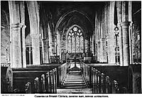 The interior of a building, symmetric with two lines of tall pillars on either side, with candelabras and wooden benches between them.