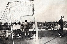 Photographie en noir et blanc d'une pelouse, d'une lucarne et de plusieurs joueurs de football.