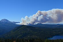A plume of brown and gray smoke rises several miles away and exits to the right of the frame, rising above rugged green conifer forest dotted with mountains