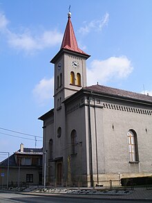 Věž kostela svatého Jiří, na kterou doprava navazuje kostelní loď. Na věži jsou umístěny hodiny a věž je zakončena červenou špičatou střechou, na jejímž vršku je kříž. Před kostelem vede k němu přístupová cesta. V pozadí za kostelem je nízký jednopodlažní dům.