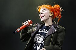 A woman with orange hair sings into a wireless orange microphone. She is wearing a black jacket, opened to show a black-and-white shirt. An earpiece can be seen in her left ear.