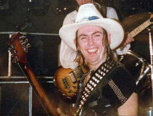 A man with shoulder-length hair wears a black t-shirt and a white cowboy hat. His guitar sling sports studs.