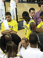 basketball players in maize uniforms have their attention on a man in a white shirt who is seated or kneeling below them. They look over his shoulders as other people look on.